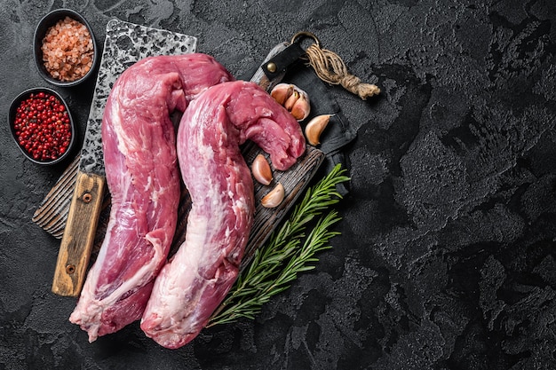 Pork tenderloin meat raw uncooked fillet on butcher board with\
meat cleaver black background top view copy space