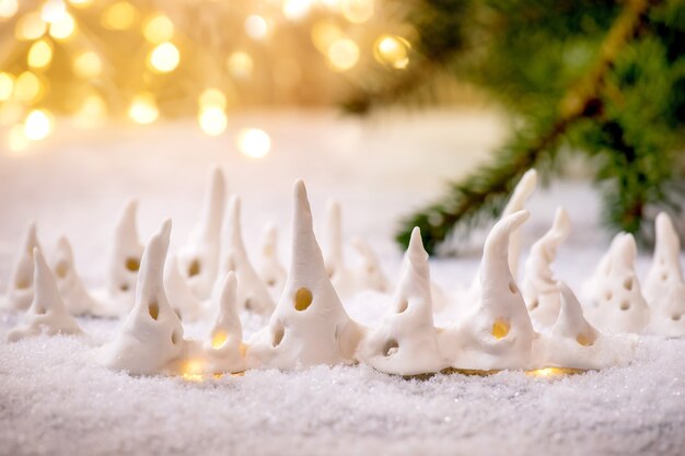 Porseleinen kersthuisjes. Set ambachtelijke handgemaakte kerstversiering huizen met gloeiende ramen op sneeuw met bokeh vakantielichten en sparren takken.