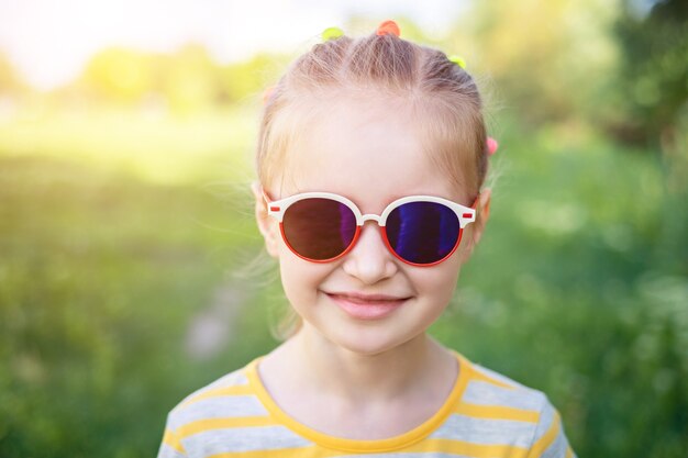 Ritratto di una bambina sorridente in graziosi occhiali da sole moderni sullo sfondo di un parco verde