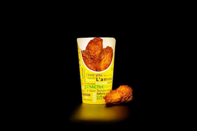 A portion of chicken wings barbecue in a cardboard Cup with inscriptions in different languages. Isolated on black background.