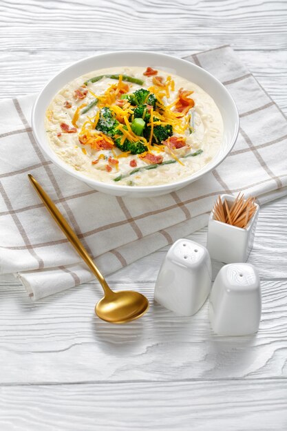 Portion of cream soup with broccoli, green bean, fried bacon, and shredded cheddar cheese in a white bowl on a wooden table