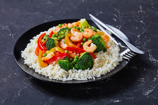 A portion of fried shrimps with bell pepper onion broccoli and green peas on steamed rice on a black plate on a concrete table landscape view from above