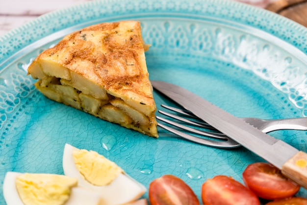 Porzione di una tipica frittata di patate o patate spagnola su un piatto azzurro con un uovo e pomodorini in un ambiente rustico cucina tradizionale spagnola fuoco selettivo sulla porzione