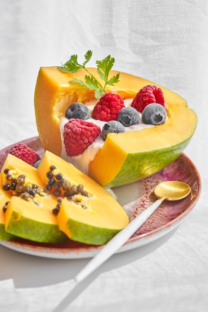 Порция вкусных фруктов папайи подается на керамической тарелке с ложкой и украшена йогуртом, малиной и черникой на белом фоне