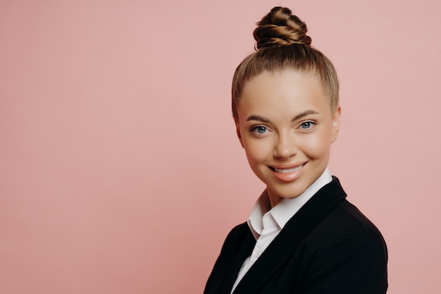 Portirat van vrolijke gelukkige vrouwelijke kantoormedewerker in donker pak met haar in knot wordt op de hoogte gebracht van haar promotie, met mooie glimlach zijwaarts kijkend naar camera, geïsoleerd over roze achtergrond