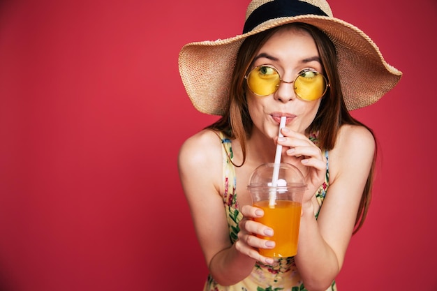 Portrair of happy and excited blond trendy woman in hat with fresh drink in hands isolated on pink background. Summer mood