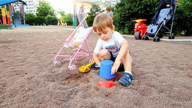 Ritratto di un bambino di 3 anni seduto nel parco giochi e che scava la sabbia con una vanga di plastica e un secchio