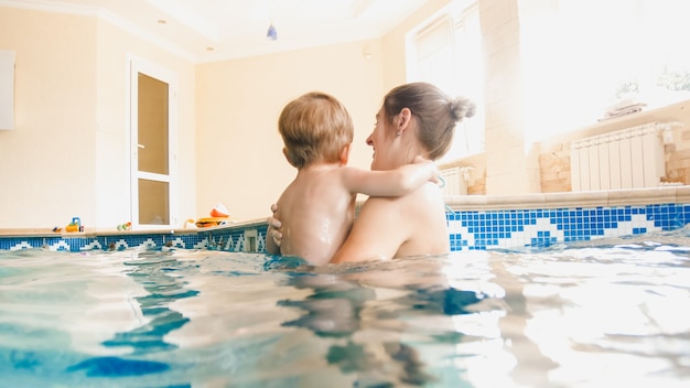 Portrait of 3 years old toddler boy with young mother swimming in the indoors pool. Child learning swimming and doing sports. Family enjoying and having fun in water