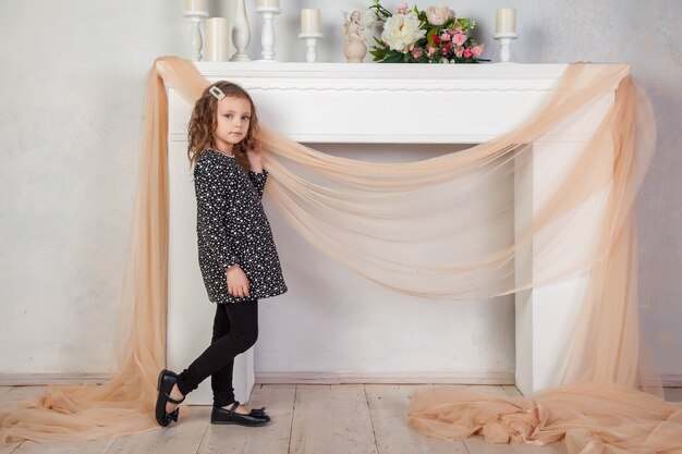 Portrait 5-6 year old girl in dress sitting by fireplace and looks at camera