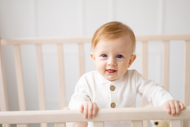 明るい子供部屋のベビーベッドに立って、綿のボディスーツで微笑んでいる生後6ヶ月の金髪の少年の肖像画子供用品のコンセプト