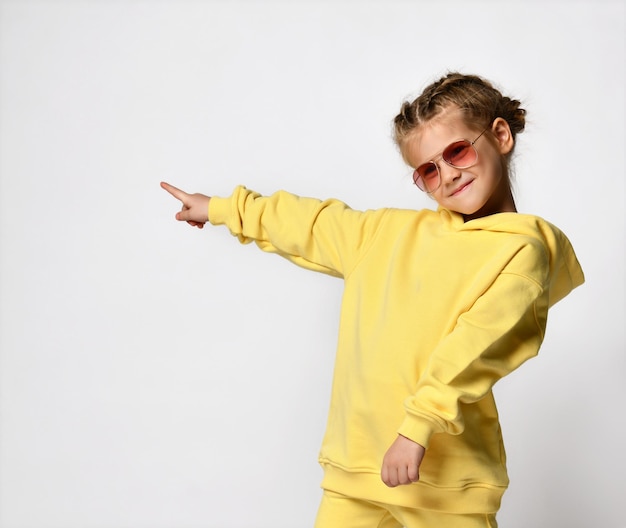 Portrait of active funny little girl pointing with fingers to the side to free space Cheerful girl dressed in a yellow tracksuit and sunglasses standing on a white background