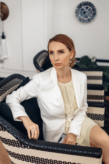 Portrait of an adorable angry boss woman in a white suit is sitting on a wicker chair with a magazine in her hands and looking into the distance. Luxury concept