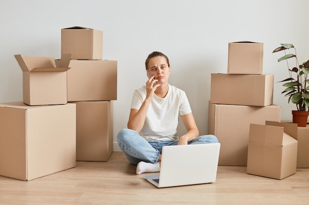 Portrait of adorable attractive woman wearing white t shirt sitting on floor surrounded with cardboard boxes with belongings and working on laptop having conversation on mobile phone