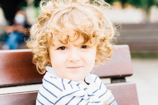 Портрет очаровательного белокурого трехлетнего мальчика с кудрями, смотрящего прямо перед собой, сидящего в парке на улице летом или весной.