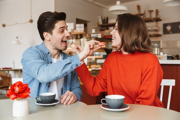 Portrait of adorable couple man and woman dating in cozy bakery, and feeding each other with macaroon biscuits