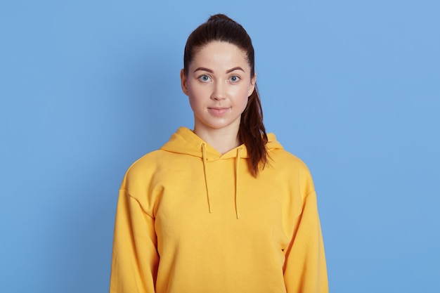 Portrait of adorable female in good mood, dark haired lady with ponytail posing isolated over blue background, wearing yellow hoodie, looks at camera.