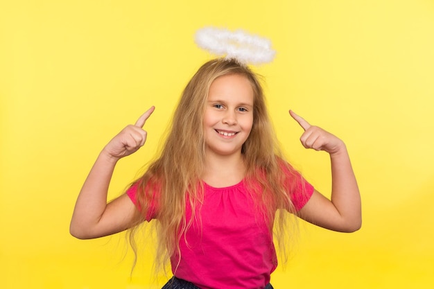 Portrait of adorable happy little girl with long fair hair pointing at angelic halo over head looking at camera with toothy smile carefree optimistic childish view indoor studio shot isolated