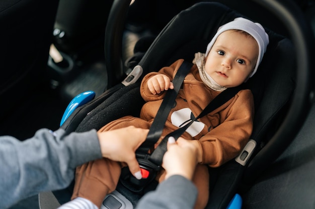 Portrait of adorable infant baby boy which unrecognizable young mother fastening safety belt in car