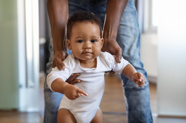 Photo portrait of adorable little black baby walking with father39s help in room