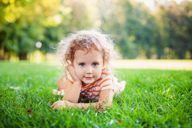 Portrait of adorable little curle girl lying on grass and propping up her face at summer green park