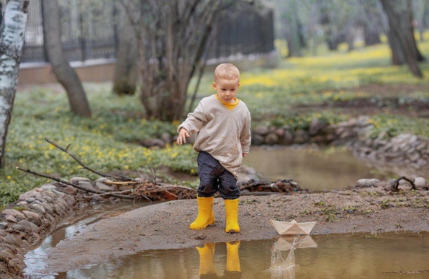 ゴム長靴の愛らしい小さな男の子の肖像画は、春の折り紙の水たまりに紙のボートを起動します