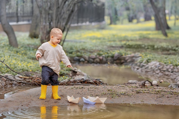 Portrait of adorable little kid boy in rubber boots launch paper boats in a puddle in spring Origami