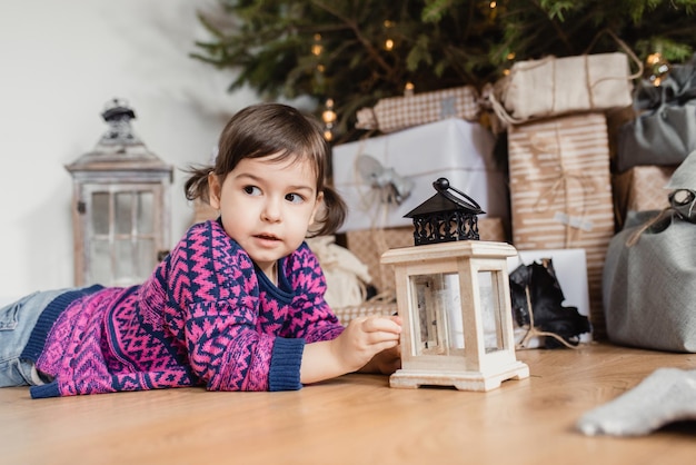 Portrait of adorable little toddler girl playing with ornaments under decorated Christmas tree on the floor in the room Merry Christmas and Happy Holidays
