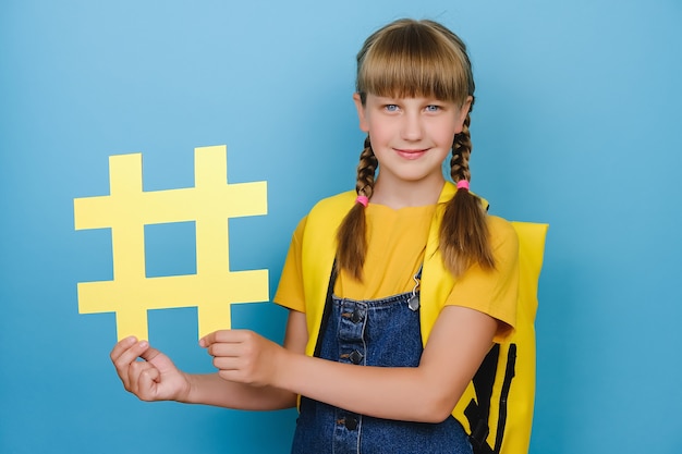 Portrait of adorable schoolgirl with yellow backpack holding big hashtag symbol and looking at camera, showing hash sign, posing over blue background. Concept of trendy social media posts and school