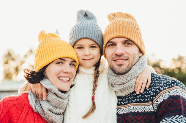 Portrait of adorable small girl wears knitted hat and sweater stands between parents, embrace them