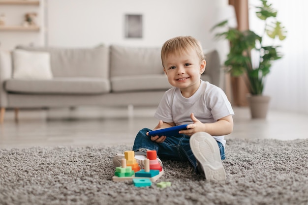 Portrait of adorable toddler boy holding smartphone sitting on floor carpet looking and smiling at