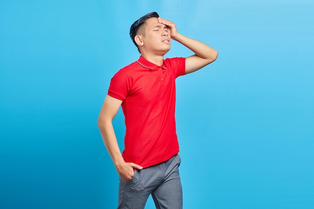 고통스러운 편두통 제스처를 받고 파란색 배경에 고립 된 머리를 들고 빨간 셔츠에 성인 아시아 남자의 초상화