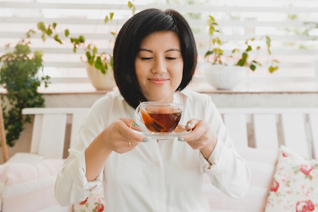 家のテラスでガラスのカップでお茶を飲むアジアの大人の女性のポートレート