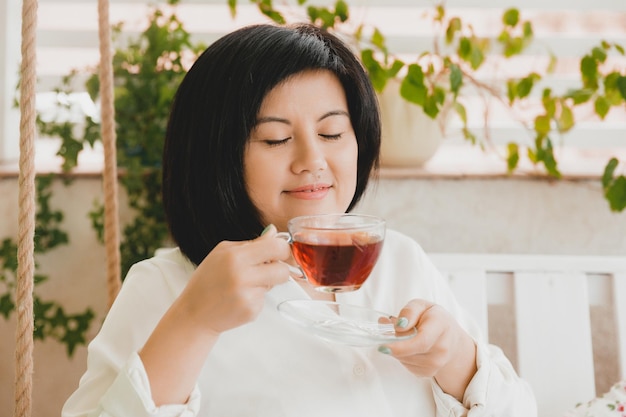 Portrait of an adult Asian woman drinking tea in a glass cup on the terrace of her house