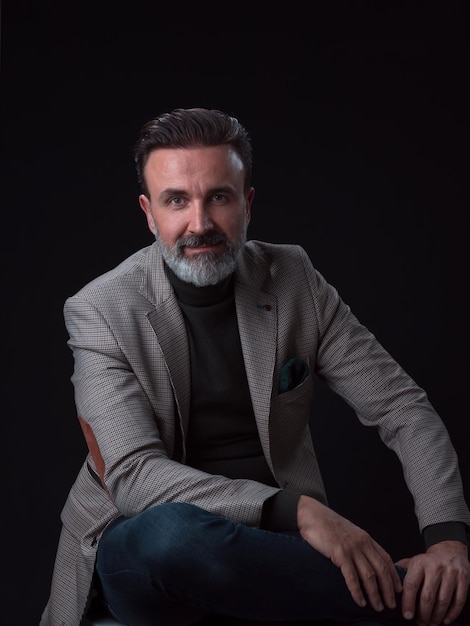 Portrait of adult businessman wearing trendy suit and sitting in modern studio on stylish chair agai