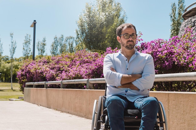 Foto ritratto di adulto latino ispanico caucasico, con barba e occhiali, serio su una sedia a rotelle guardando la telecamera, all'aperto in un bel parco