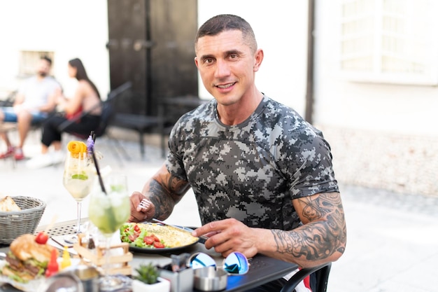 Portrait of Adult Man Eating Omelet or Scrambled Eggs Outdoors at Restaurant and Drinking Cocktail