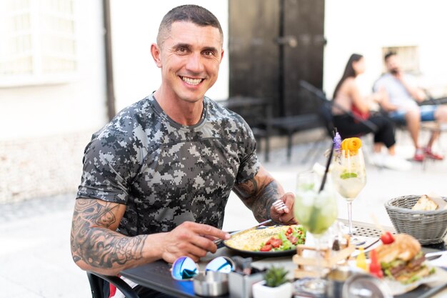 Portrait of Adult Man Eating Omelet or Scrambled Eggs Outdoors at Restaurant and Drinking Cocktail