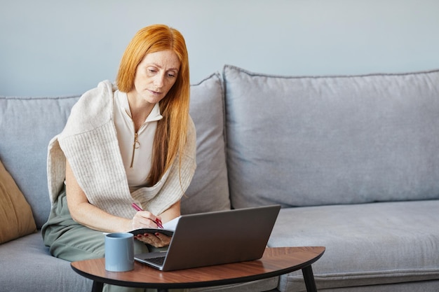 Portrait of adult red haired woman using laptop on sofa while studying online from home copy space