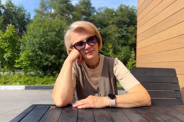 Portrait of adult senior woman sitting at table on terrace at summer sunny day in sunglasses