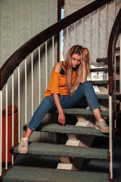 Portrait adult woman actress posing on home staircase dressed styling clothes in vintage living room. Female with long hair in style orange jacket. Concept retro fashion and outdated trend vogue