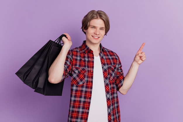 Portrait of adviser cheerful guy direct finger blank space carry store bags on violet background