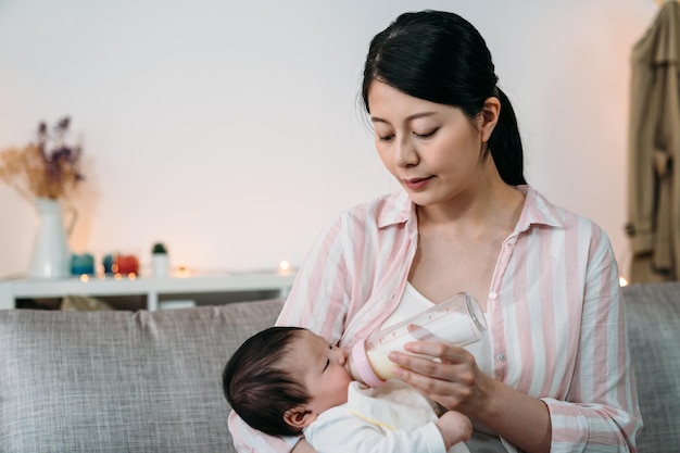 肖像画の愛情深い日本人の母親は、自宅のリビングルームのソファで乳児の哺乳瓶のミルクを手に持って食べさせています.