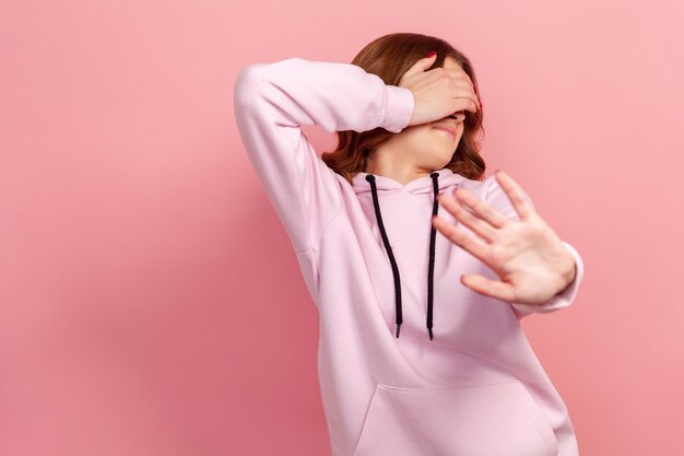 Ritratto di giovane donna timida o impaurita con i capelli ricci in felpa con cappuccio che chiude gli occhi con il palmo e mostra il gesto della mano di arresto colpo in studio al coperto isolato su sfondo rosa