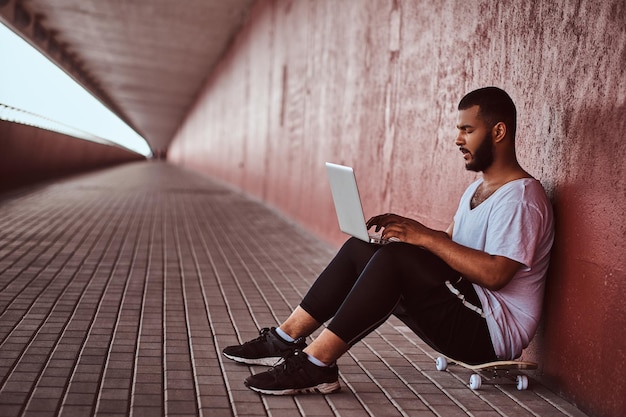 Foto ritratto di un ragazzo barbuto afroamericano vestito con una camicia bianca e pantaloncini sportivi che lavora su un laptop mentre è seduto su uno skateboard sotto il ponte.