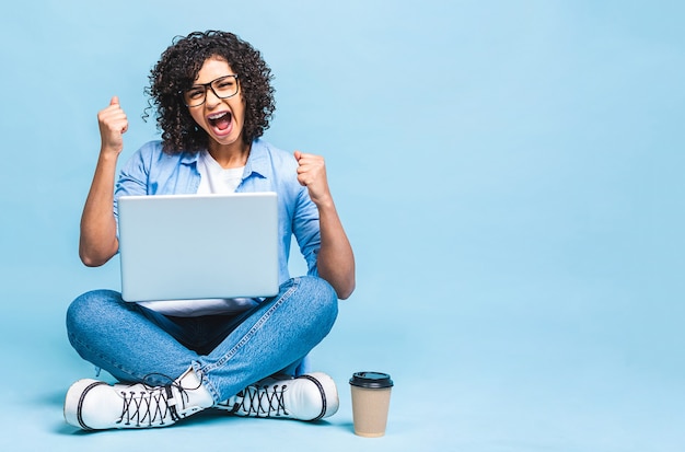 Portrait of african american black woman in casual sitting on floor in lotus pose and holding laptop isolated over blue background