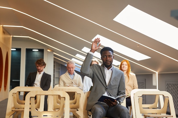 Portrait of African-American businessman raising hand to ask question while sitting in audience of contemporary conference hall interior, copy space