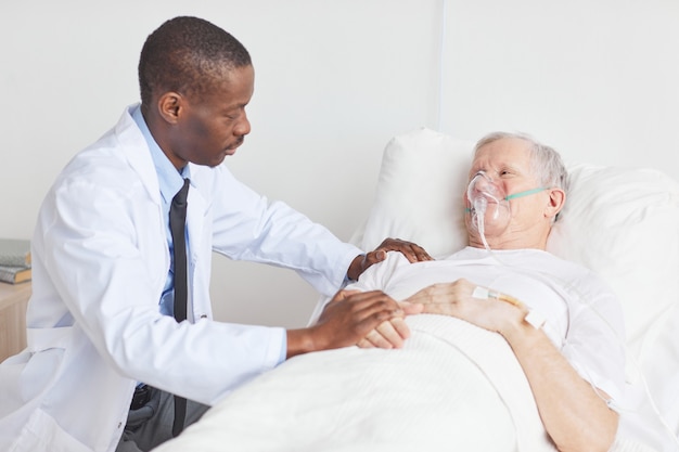 Portrait of African-American doctor caring for senior man lying in hospital bed with oxygen supplementation mask, copy space