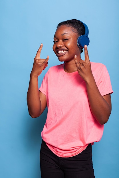 Portrait of african american funny teenager with headphones enjoying rock music playlist dancing alone in studio with blue background. Smiling student having fun listening energetic song