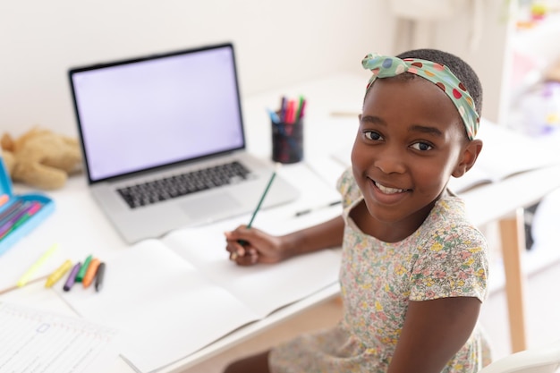 オンライン学校レッスン用のコピースペース付きラップトップを使用して机にいるアフリカ系アメリカ人の女の子のポートレート