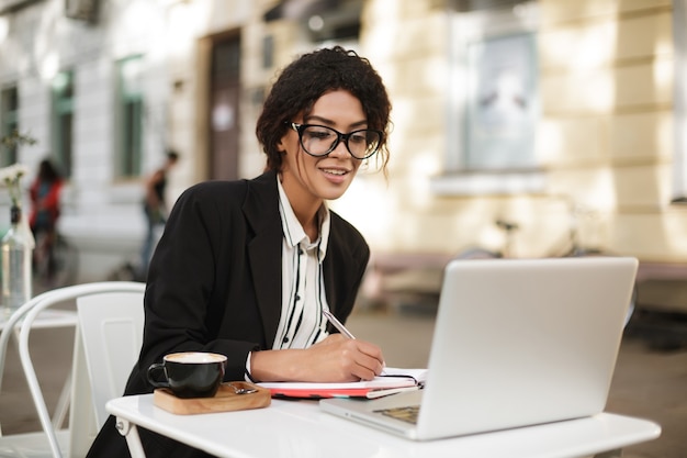 Portrait of African American girl sitting at the table of cafe and writing in her notebook while 